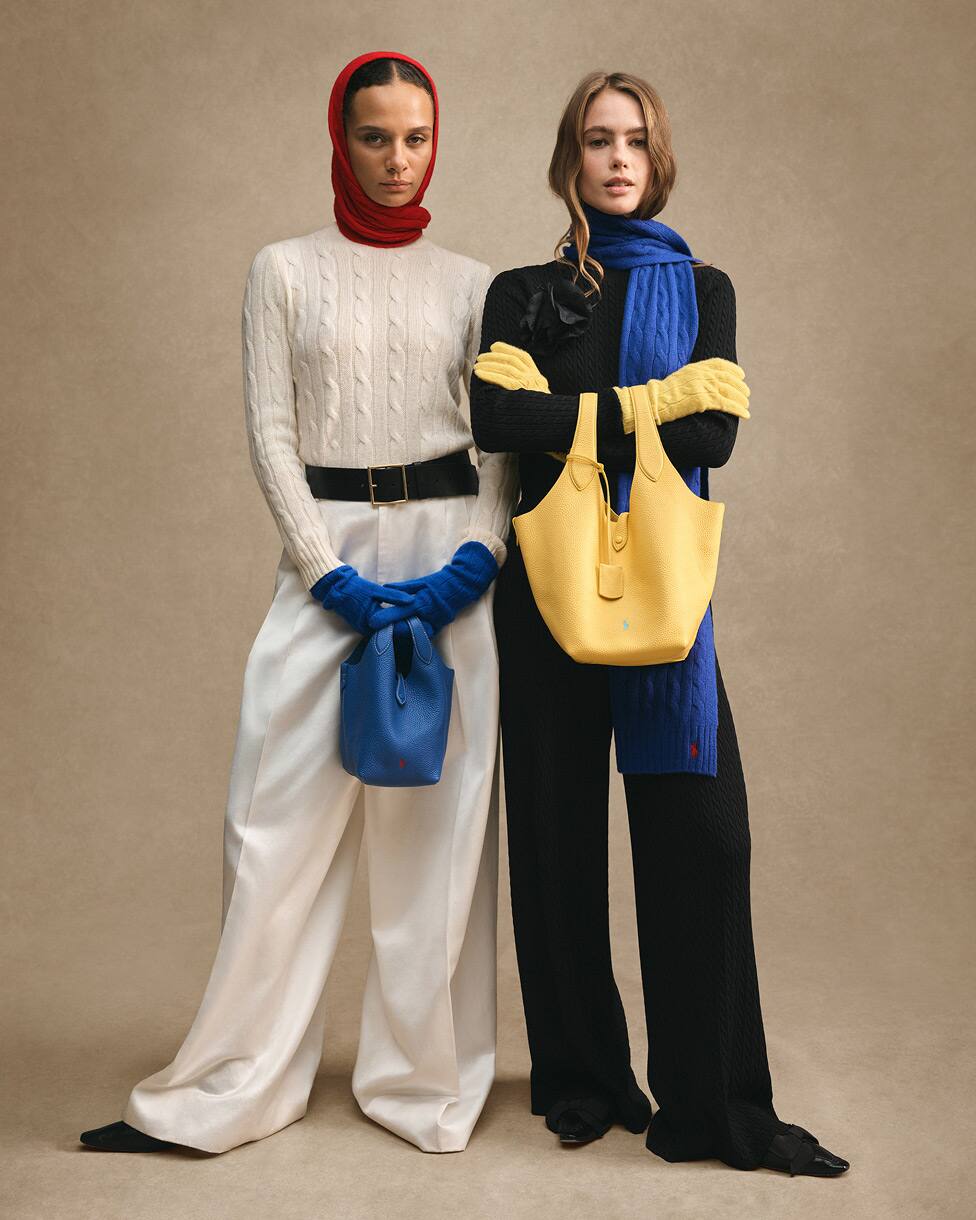 Zwei stehende Frauen, eine in einem cremefarbenen Stricklook mit roter Kopfbedeckung und blauen Handschuhen, die andere in einem schwarzen Stricklook mit blauem Schal und gelber Tasche.
