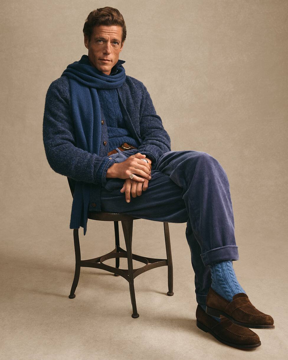 Sitzender Mann in blauem Stricklook mit passendem Schal und braunen Loafern.
