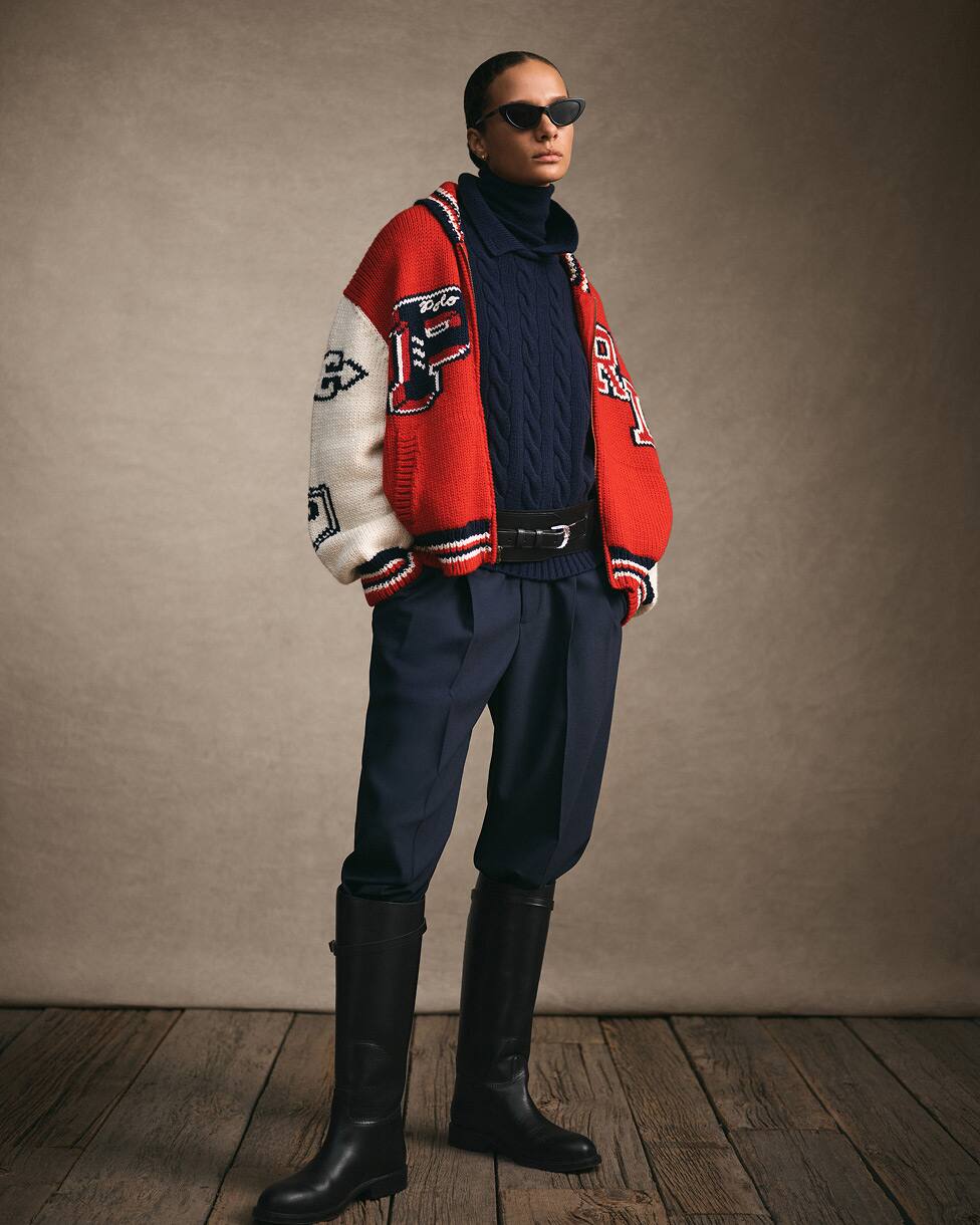 Stehende Frau in rot-weißer College-Jacke mit marineblauem Pullover und marineblauer Hose sowie hohen schwarzen Stiefeln.