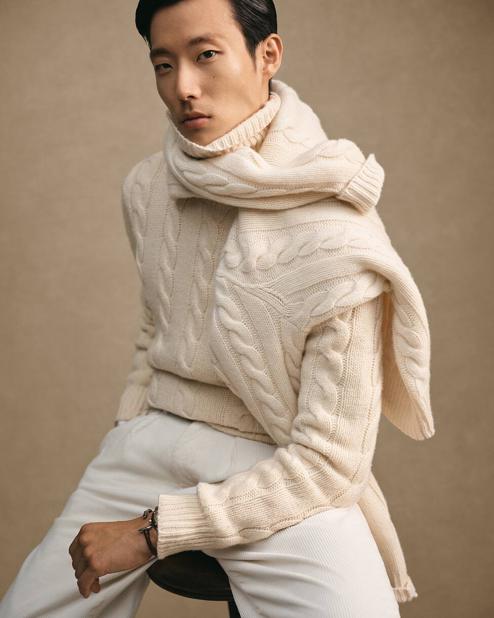 Man seated wearing a cream cable knit jumper, matching scarf and white trousers.