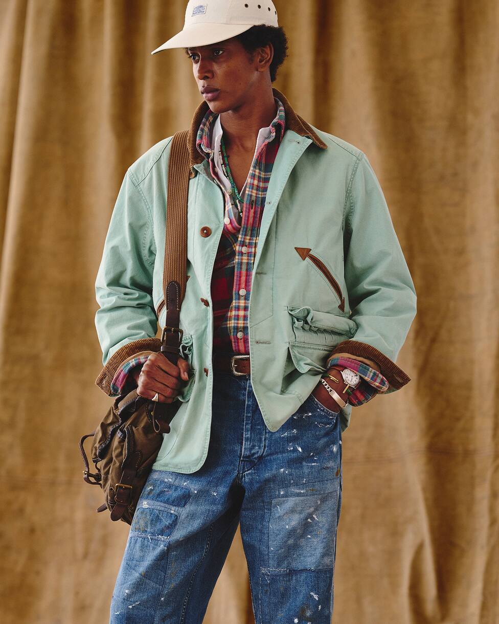 Man wearing a green jacket over a plaid shirt with blue jeans, a cream cap, and a bag.